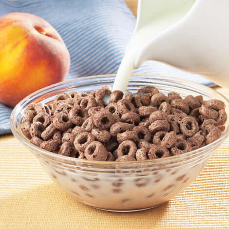 Bowl of protein cereal with milk pouring over, alongside a peach on a blue and yellow tablecloth.
